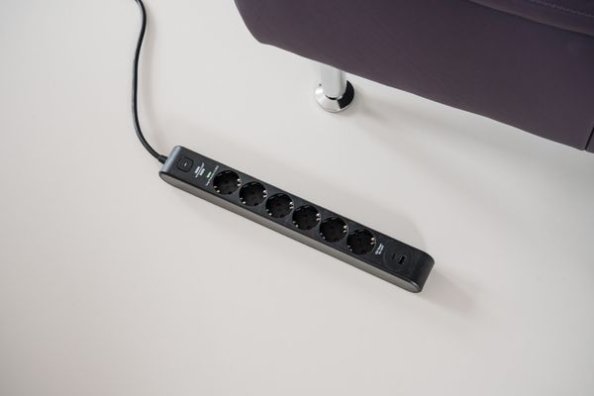Schwarze Brennenstuhl Steckdosenleiste mit mehreren Steckdosen und USB-Anschlüssen auf dem Boden neben einem Sofa.